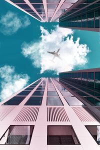 A plane flying over tall skyscrapers with a clear sky and clouds.