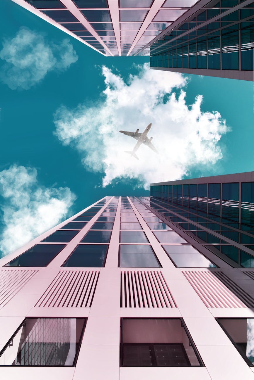 A plane flying over tall skyscrapers with a clear sky and clouds.